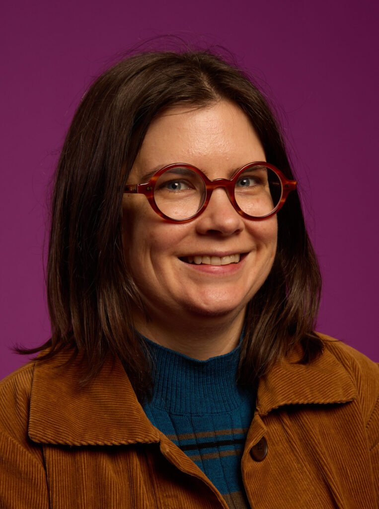 A woman smiling, wearing glasses, a brown jacket and a blue shirt